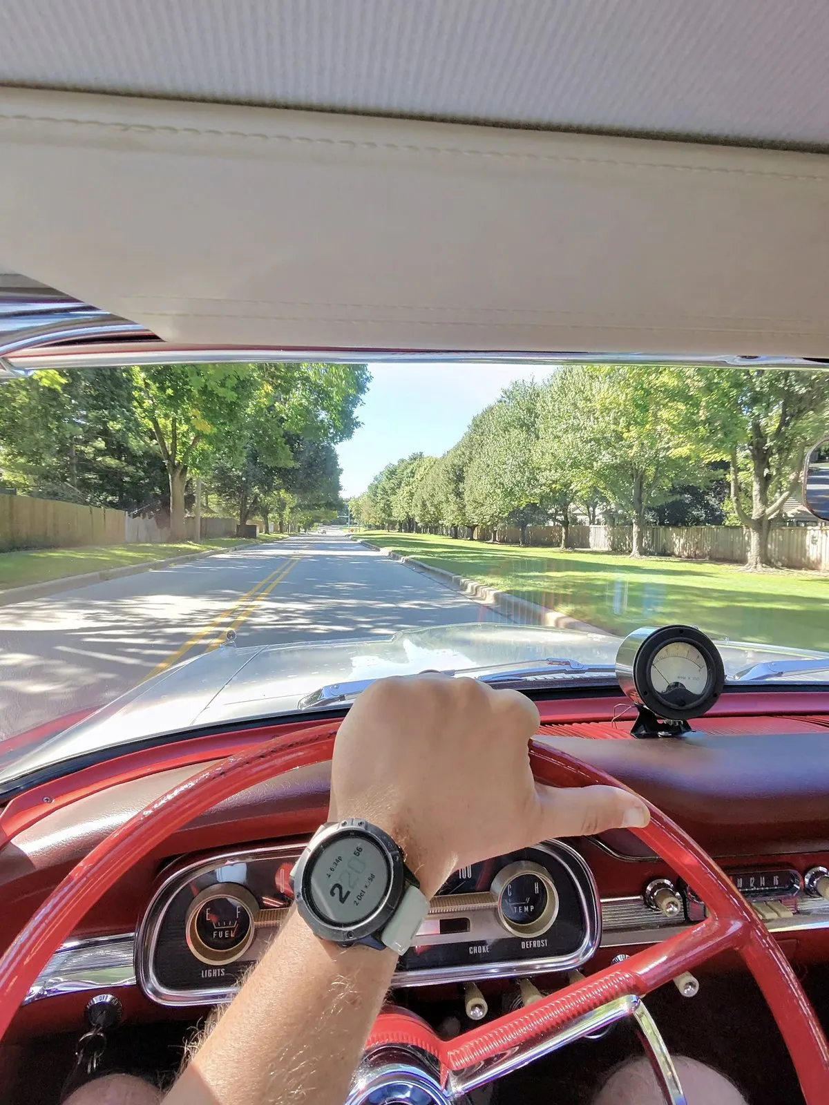 Driver view over the red steering wheel of a 1963 Ford Falcon, looking down a sunny tree-lined street
