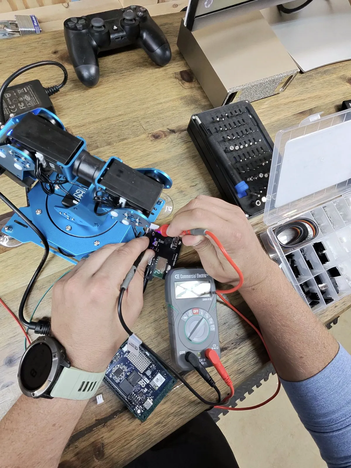 Hands holding multimeter probes on the stock controller board of the Hiwonder xArm, with the meter on the workbench and an Arduino Uno Q waiting in the foreground